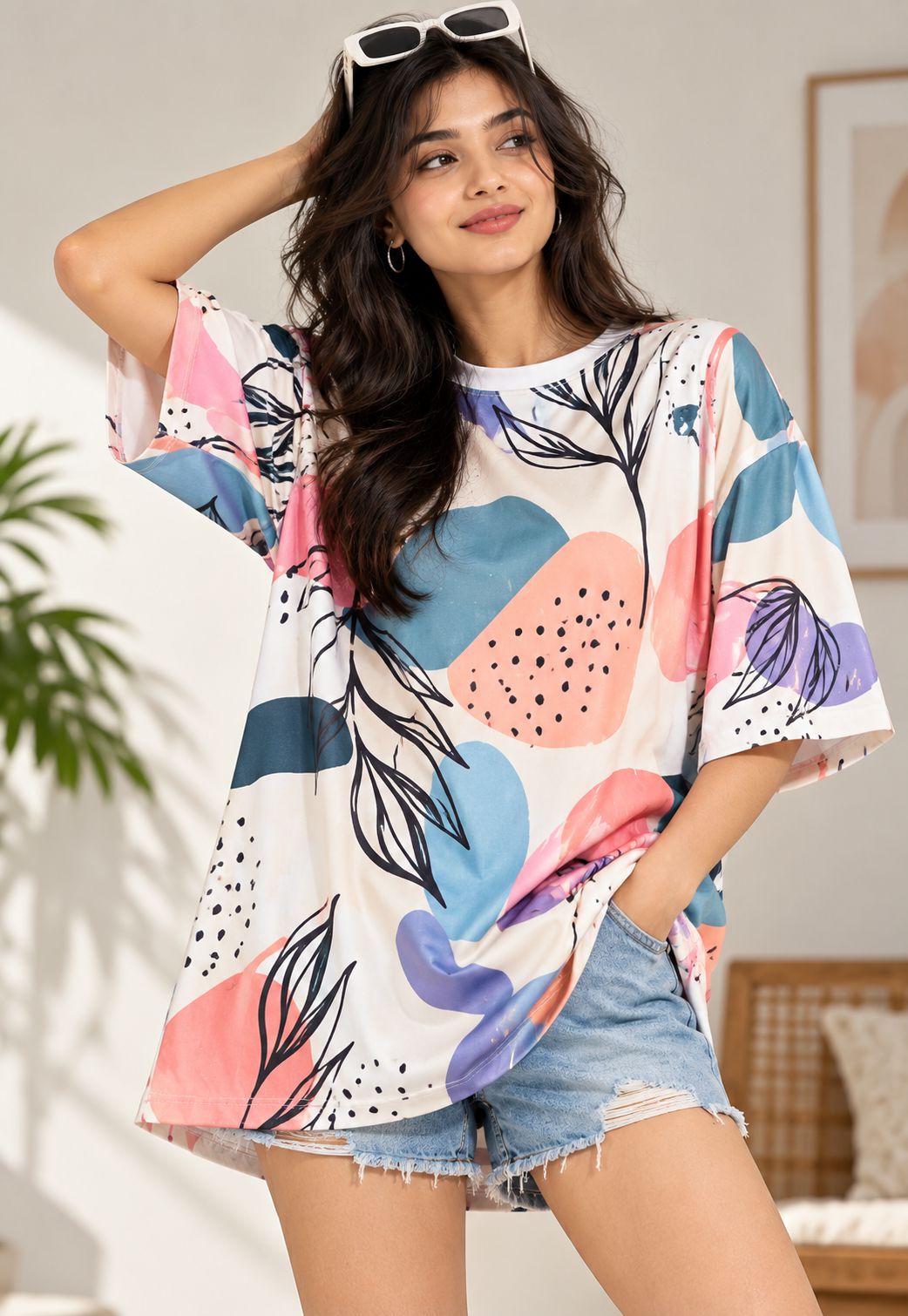 A woman wearing a loose white tunic top with a colorful abstract print in pink, blue, purple, and teal, paired with denim shorts in a bright indoor setting.