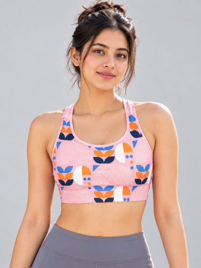 A woman wearing a pink sports bra with a colorful geometric pattern of orange, navy blue, and white shapes, paired with gray athletic leggings, set against a solid gray studio background.
