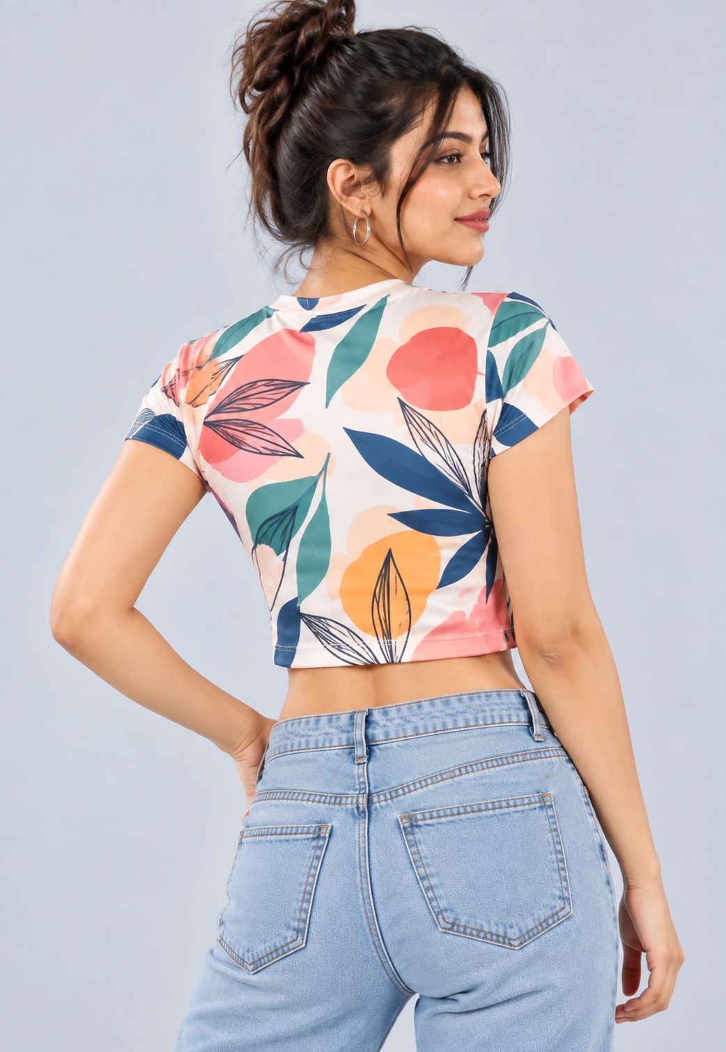 A woman wearing a short-sleeve crop top with a colorful floral pattern, shown from the back. She is wearing light blue jeans against a solid light gray studio background.