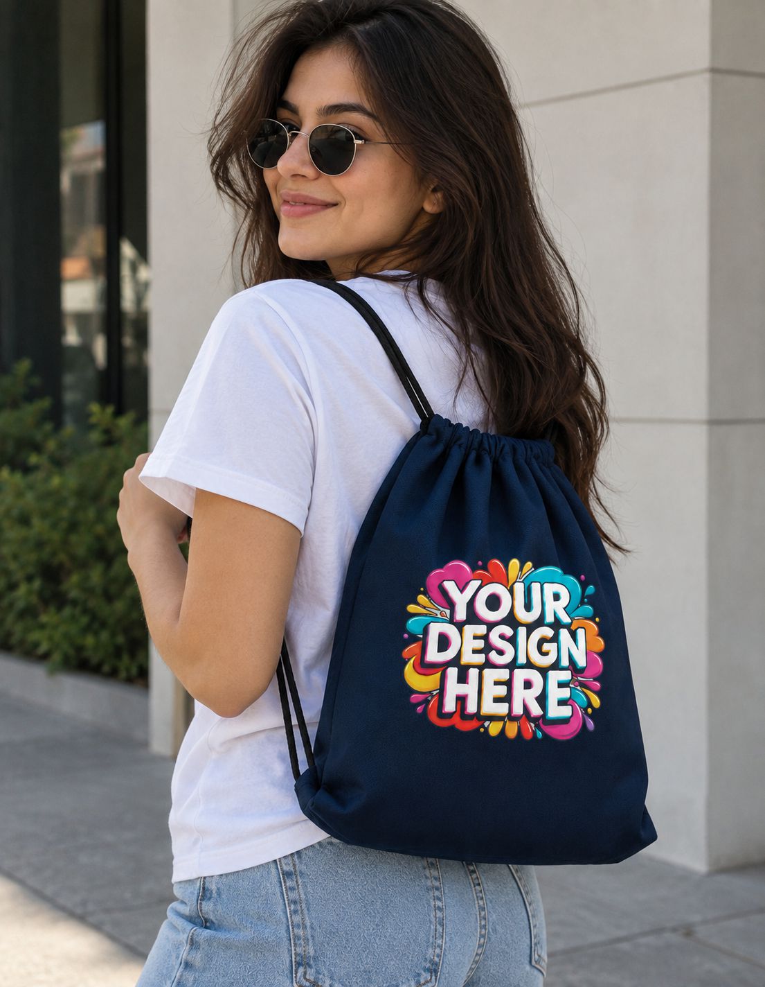 A woman wearing round sunglasses and a white t-shirt carries a navy blue drawstring backpack with a colorful 'YOUR DESIGN HERE' mockup graphic on the back, set against a modern building exterior.