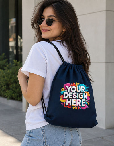 A woman wearing round sunglasses and a white t-shirt carries a navy blue drawstring backpack with a colorful 'YOUR DESIGN HERE' mockup graphic on the back, set against a modern building exterior.