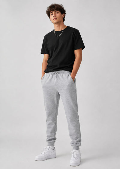 A young man modeling a black crewneck t-shirt, light gray heather sweatpants, and white sneakers against a light gray studio background.