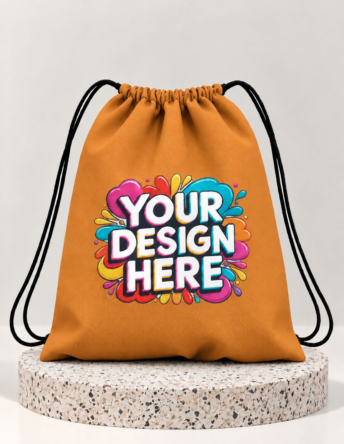 An orange drawstring bag with black cords, featuring a colorful 'YOUR DESIGN HERE' graphic on the front, displayed on a round speckled stone pedestal against a light gray background.