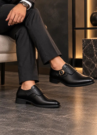 Black faux leather monk strap shoes with woven panels and gold-tone buckles, worn with trousers