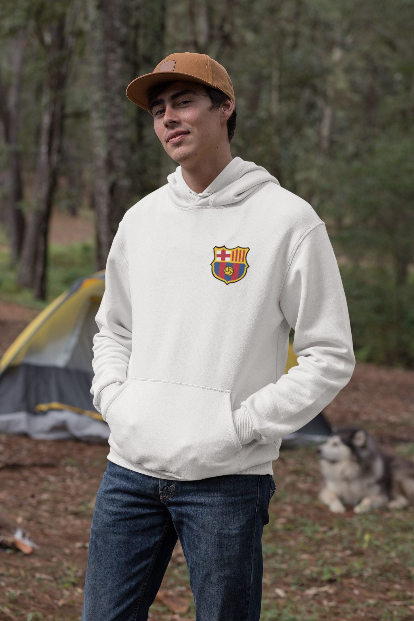 Person wearing a white hoodie with a logo, standing outdoors with trees and a tent in the background.