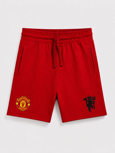 Red Manchester United athletic shorts with a drawstring waist, side pockets, and embroidered club crests on the left and right legs, shown on a light gray background.