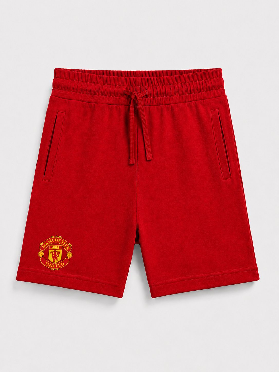 Red Manchester United sweat shorts with an elastic drawstring waist and a yellow embroidered club crest on the left leg, shown against a light gray background.