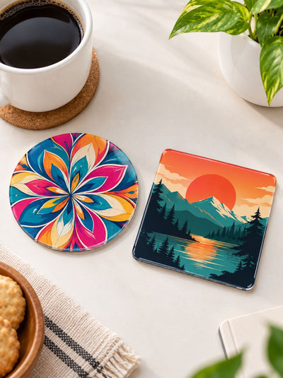 Two decorative coasters on a light tabletop: one round coaster with a bright multicolor floral mandala pattern and one square coaster with a mountain lake sunset illustration. A cup of black coffee, plant leaves, and a small bowl of cookies are partially visible around them.