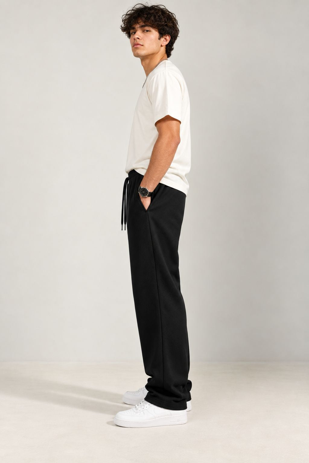 A man wearing a white t-shirt, black drawstring sweatpants, and white sneakers, standing in profile against a light gray studio background.