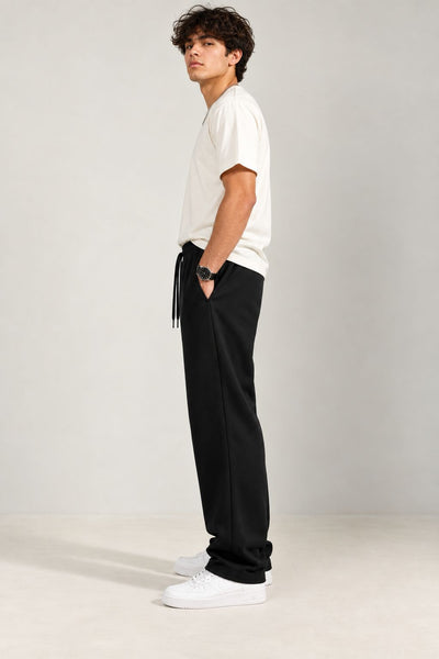 A man wearing a white t-shirt, black drawstring sweatpants, and white sneakers, standing in profile against a light gray studio background.