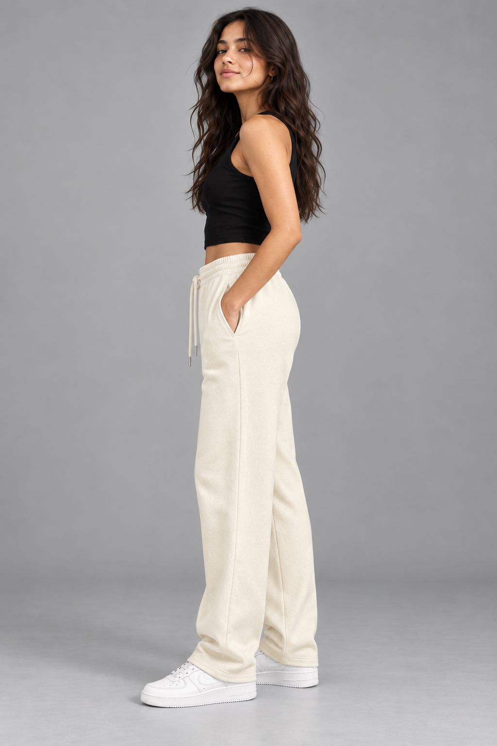 A woman modeling a pair of cream-colored, wide-leg sweatpants with a drawstring waist, paired with a black crop top and white sneakers, set against a solid gray studio background.