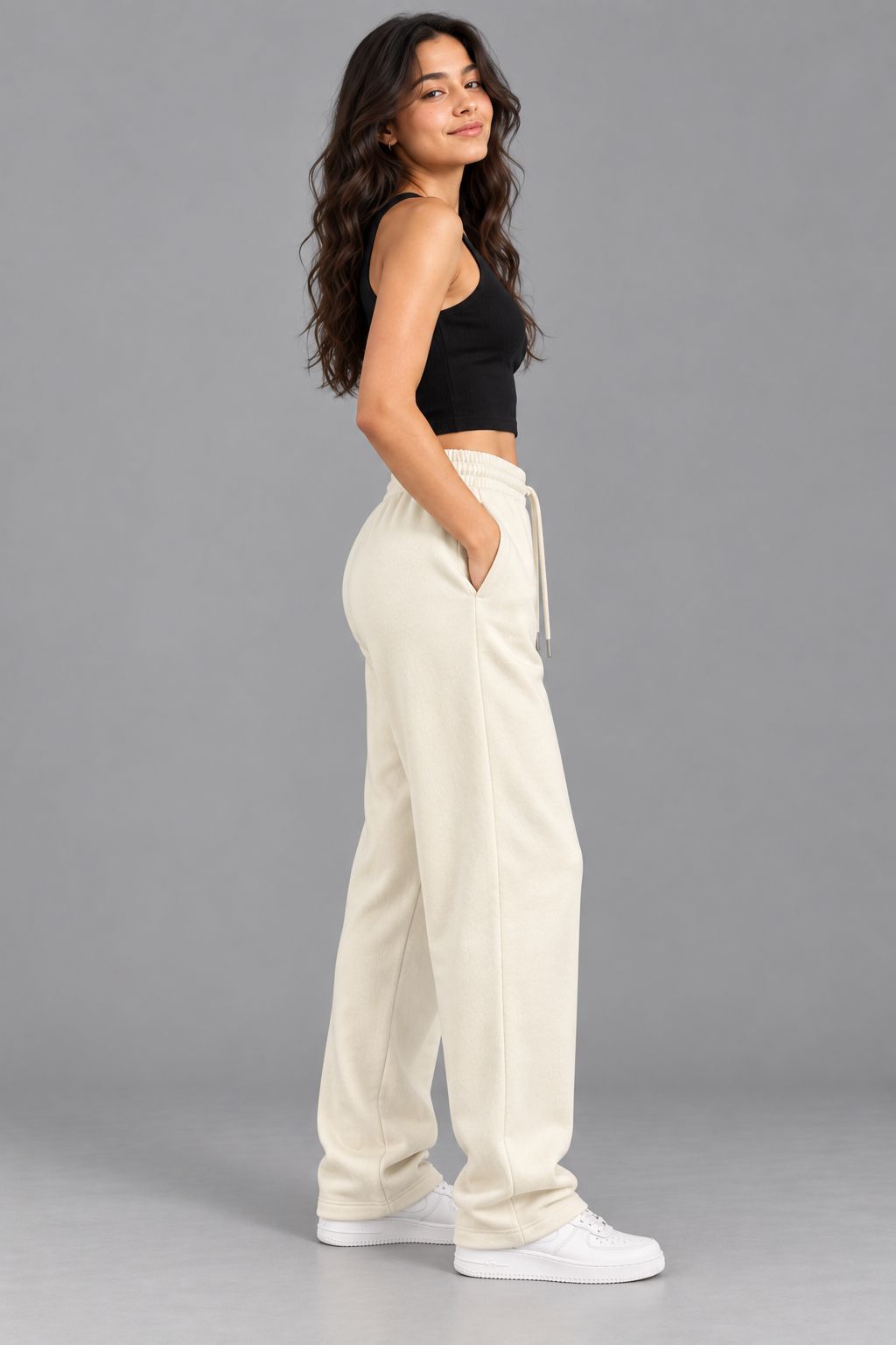 A woman modeling a pair of cream-colored, wide-leg sweatpants with a drawstring waist, paired with a black crop top and white sneakers, set against a solid gray studio background.