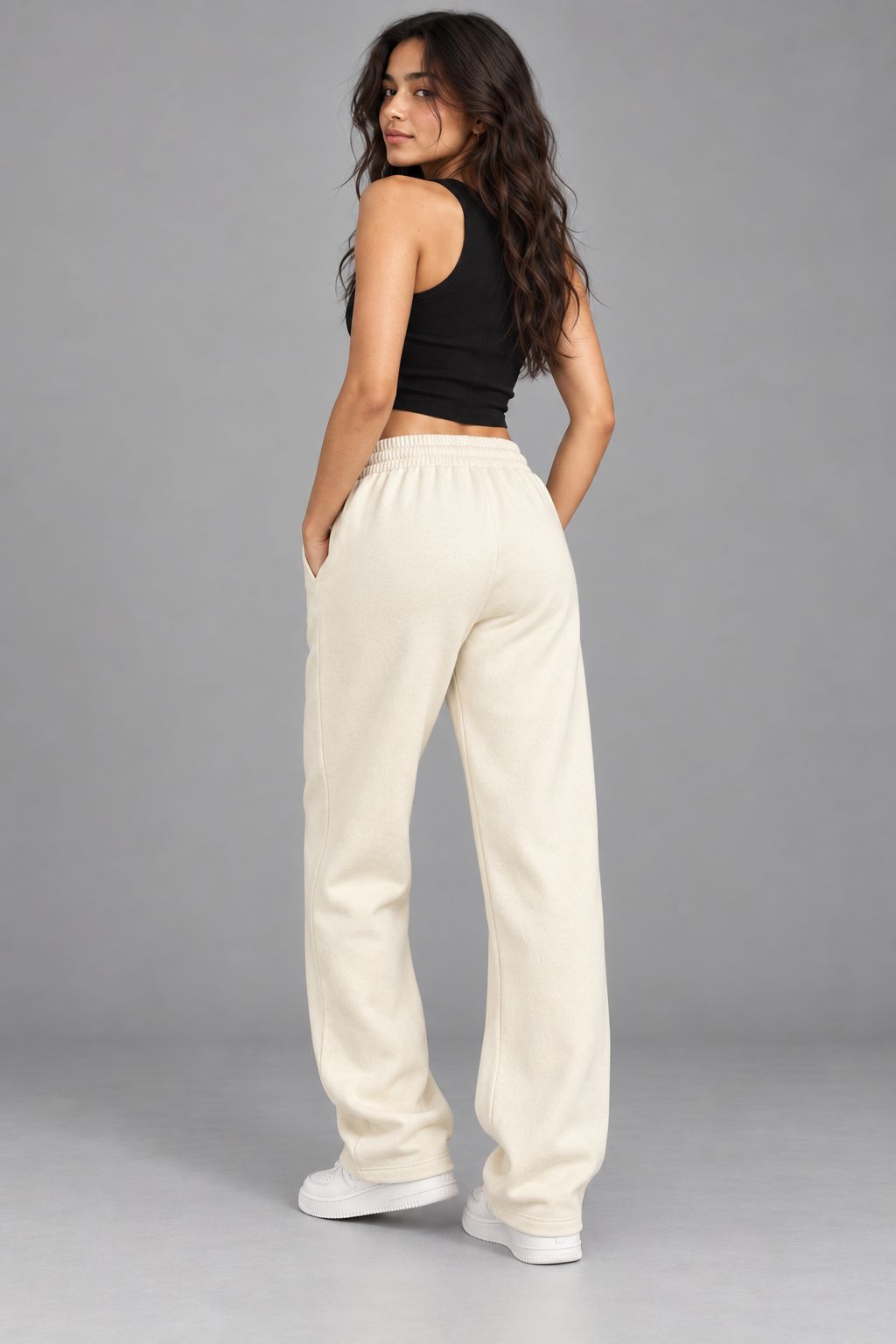 A woman modeling a pair of cream-colored, wide-leg sweatpants with a matching elastic waistband, paired with a black crop top and white sneakers, shown from a rear three-quarter angle against a solid gray studio background.